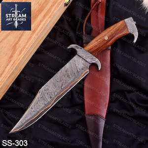 May include: A Damascus steel hunting knife with a wooden handle and a leather sheath. The blade has a unique pattern, and the handle features a decorative pin. The knife is labeled "SS-303" and is displayed on a black surface.