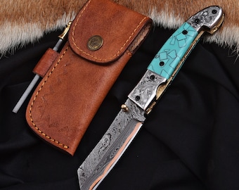 May include: A folding knife with a turquoise handle and a patterned blade, next to a brown leather sheath. The knife has a silver bolster with decorative etching. A sharpening rod is included with the sheath. The text "SS-324" is visible.