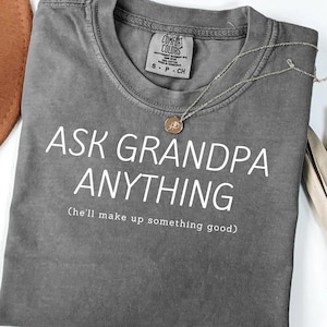 May include: A gray Comfort Colors t-shirt with the text "ASK GRANDPA ANYTHING (he'll make up something good)" in white. A gold necklace with a pendant rests on the shirt. The shirt is size S-P-CH.
