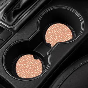 May include: Two car coasters with a peach-colored background and a pattern of small white dots. The coasters are placed inside the cup holders of a black car interior. The car interior is made of black leather and plastic.