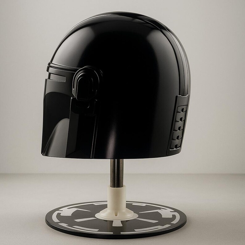May include: A black, glossy helmet, similar to a Mandalorian helmet, is presented on a stand. The helmet has a smooth, curved design with detailed side panels. The stand has a black base with a white emblem.