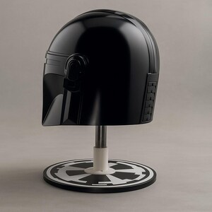 May include: A black helmet, similar to a Mandalorian design, is presented on a stand. The helmet has a smooth, curved form with detailed side panels and a visor. The stand includes a white base and a black and white emblem.