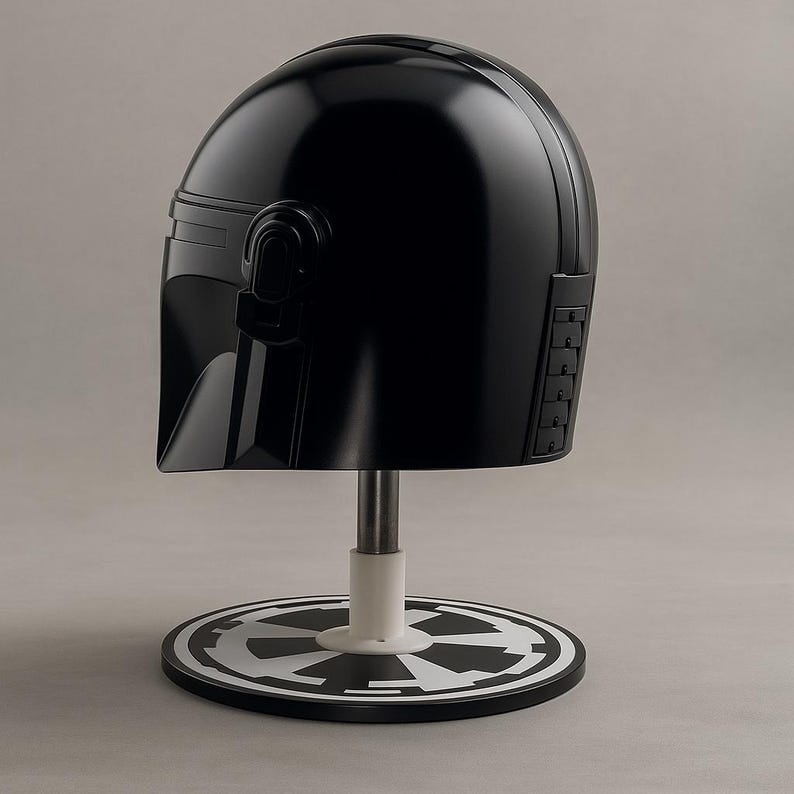 May include: A black helmet, similar to a Mandalorian design, is presented on a stand. The helmet has a smooth, curved form with detailed side panels and a visor. The stand includes a white base and a black and white emblem.