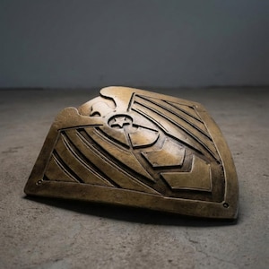 Pode incluir: Um escudo de cor bronze com um design em relevo apresentando uma &aacute;guia, uma estrela e formas geom&eacute;tricas. O escudo tem uma forma curva e est&aacute; apoiado em uma superf&iacute;cie de concreto. O design sugere um tema hist&oacute;rico ou de fantasia.