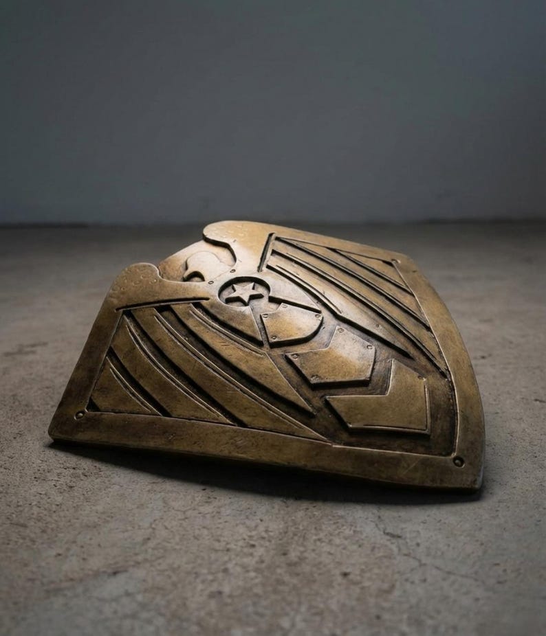 Pode incluir: Um escudo de cor bronze com um design em relevo apresentando uma &aacute;guia, uma estrela e formas geom&eacute;tricas. O escudo tem uma forma curva e est&aacute; apoiado em uma superf&iacute;cie de concreto. O design sugere um tema hist&oacute;rico ou de fantasia.