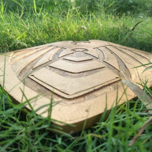Pode incluir: Um escudo dourado com um design geom&eacute;trico detalhado, repousando na grama verde. O escudo tem uma superf&iacute;cie texturizada e um padr&atilde;o sim&eacute;trico, provavelmente para fins decorativos ou cosplay.
