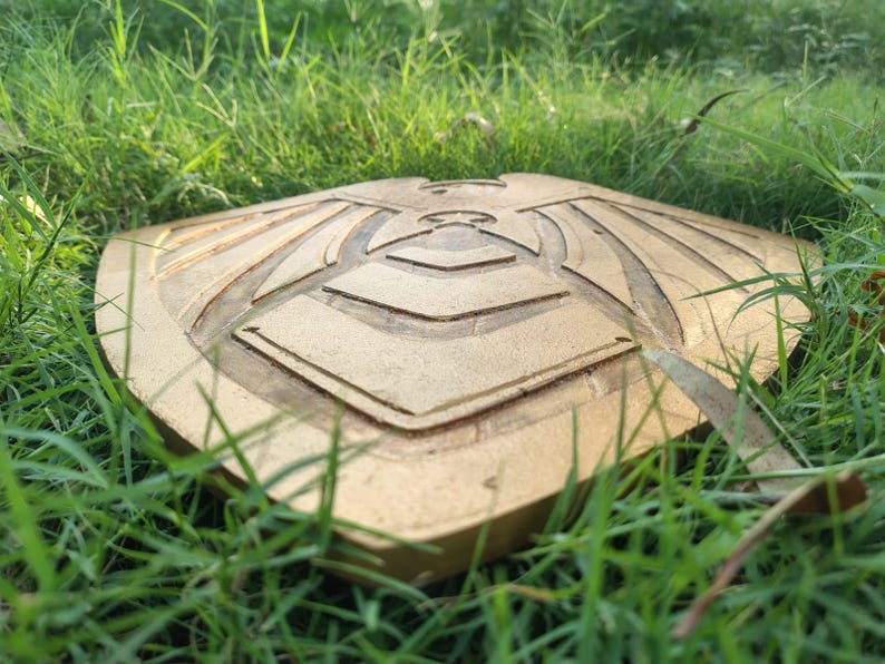 Pode incluir: Um escudo dourado com um design geom&eacute;trico detalhado, repousando na grama verde. O escudo tem uma superf&iacute;cie texturizada e um padr&atilde;o sim&eacute;trico, provavelmente para fins decorativos ou cosplay.