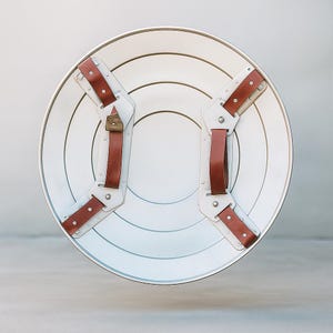 May include: A white and silver shield with brown leather straps and rivets. The shield has concentric circles and a silver rim. The straps are attached to the back of the shield with metal hardware. A classic superhero accessory.