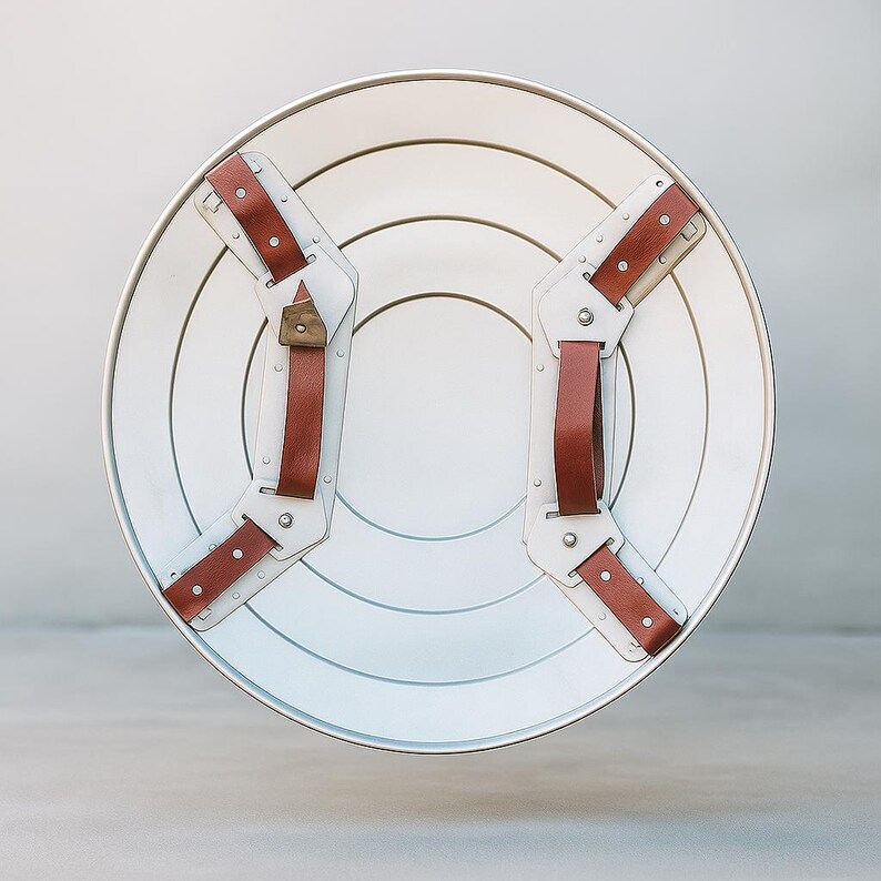 May include: A white and silver shield with brown leather straps and rivets. The shield has concentric circles and a silver rim. The straps are attached to the back of the shield with metal hardware. A classic superhero accessory.