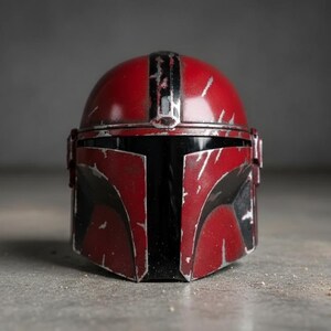 May include: A red and black Mandalorian helmet with silver accents. The helmet has a weathered appearance with scratches and wear marks. The visor is black, and the helmet is set against a gray background. A detailed replica of a popular sci-fi character's helmet.