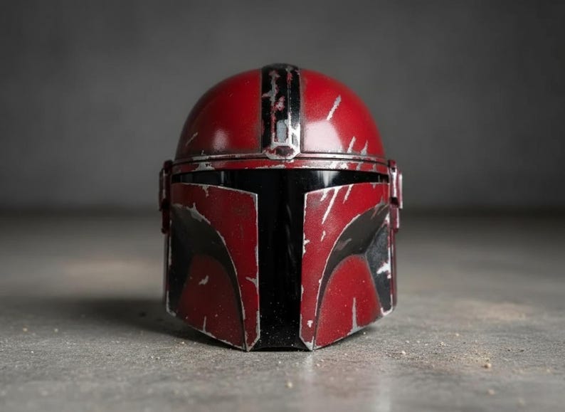 May include: A red and black Mandalorian helmet with silver accents. The helmet has a weathered appearance with scratches and wear marks. The visor is black, and the helmet is set against a gray background. A detailed replica of a popular sci-fi character's helmet.