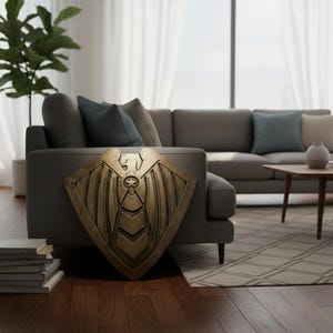 Pode incluir: Um escudo de cor bronze com um design de estrela e asas, encostado a um sof&aacute; cinza. O escudo tem uma superf&iacute;cie detalhada e texturizada e uma forma geom&eacute;trica. O fundo inclui uma sala de estar com um sof&aacute;, almofadas e uma planta.