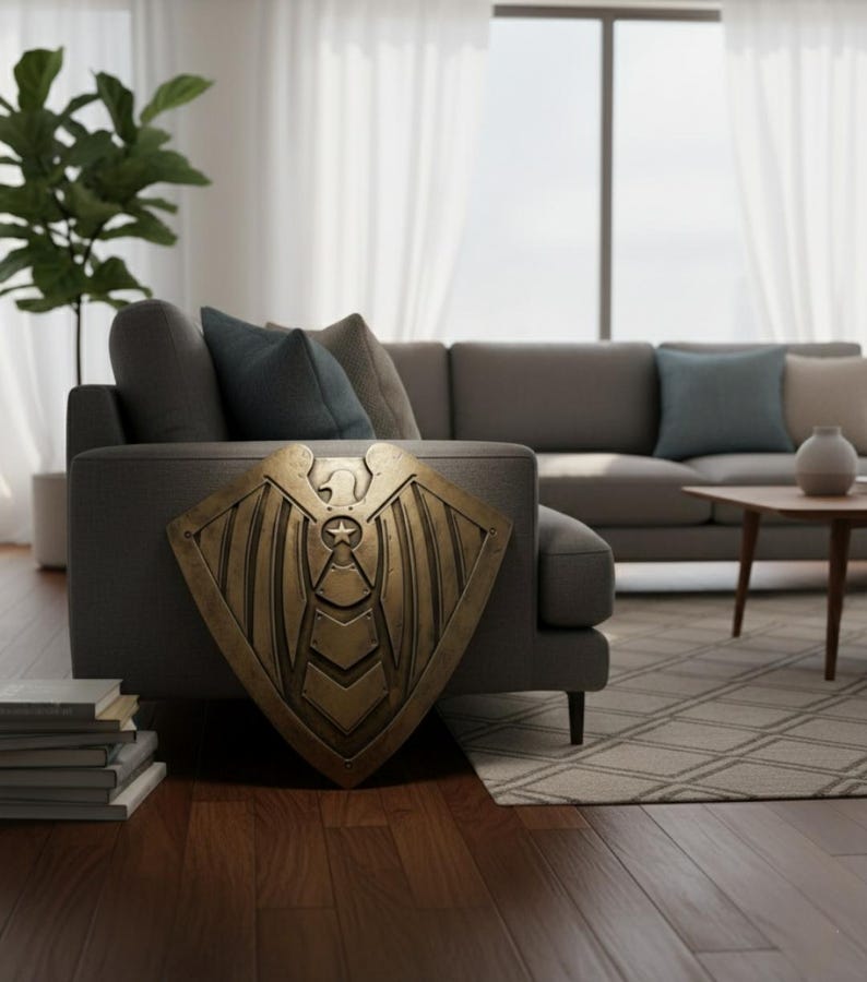 Pode incluir: Um escudo de cor bronze com um design de estrela e asas, encostado a um sof&aacute; cinza. O escudo tem uma superf&iacute;cie detalhada e texturizada e uma forma geom&eacute;trica. O fundo inclui uma sala de estar com um sof&aacute;, almofadas e uma planta.