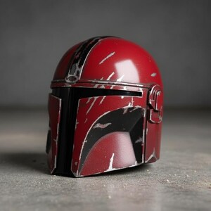 May include: A red and black Mandalorian helmet with silver accents. The helmet has a distressed, battle-worn appearance with scratches and wear marks. The helmet is a replica of the one worn by the character in the Star Wars series.