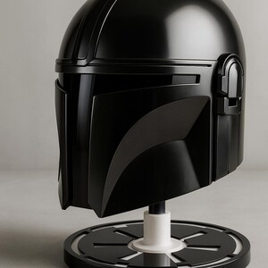 May include: A black Mandalorian helmet replica, presented on a black and white stand. The helmet has a glossy finish and detailed design, showcasing the iconic shape and visor.