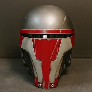 Może przedstawiać: Szczegółowe zbliżenie hełmu, głównie szarego z czerwonymi detalami. Hełm ma zaokrągloną koronę i osłonę twarzy z kanciastymi elementami. Projekt zawiera widoczne ślady zużycia, sugerujące estetykę zniszczoną przez walkę.