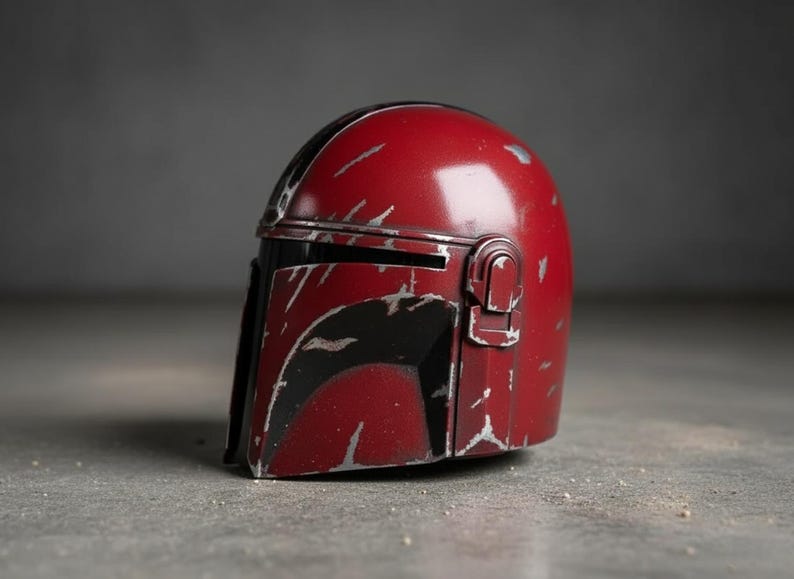 May include: A red Mandalorian helmet with black accents and silver weathering details. The helmet features a visor and a distinctive T-shaped opening. This collectible item is a replica of the iconic helmet from the Star Wars series.