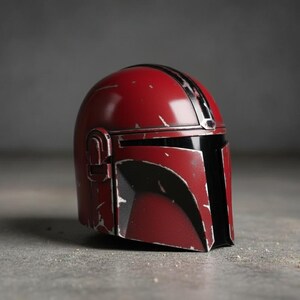 May include: A red and black Mandalorian helmet with a weathered appearance. The helmet features a glossy finish with black accents and a vertical stripe. The design includes a visor and side details, set against a neutral background.