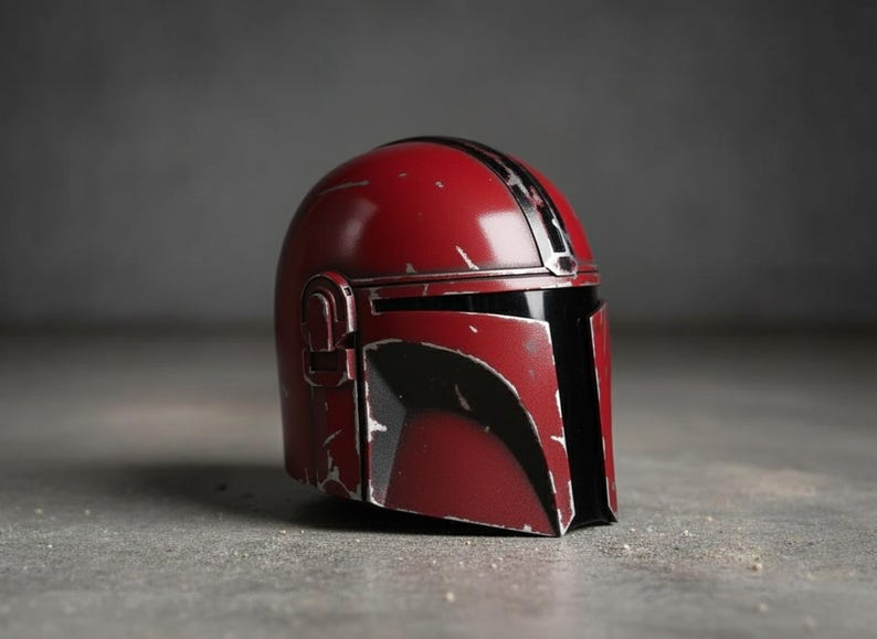 May include: A red and black Mandalorian helmet with a weathered appearance. The helmet features a glossy finish with black accents and a vertical stripe. The design includes a visor and side details, set against a neutral background.