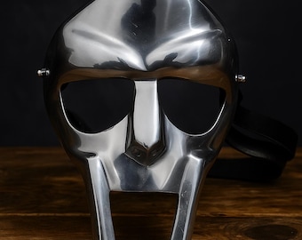 May include: A polished silver metal mask with a distinctive, angular design. The mask features large eye openings, a prominent nose piece, and a mouth-like opening. A black strap is attached to the sides. The mask is likely for costume or decorative purposes.