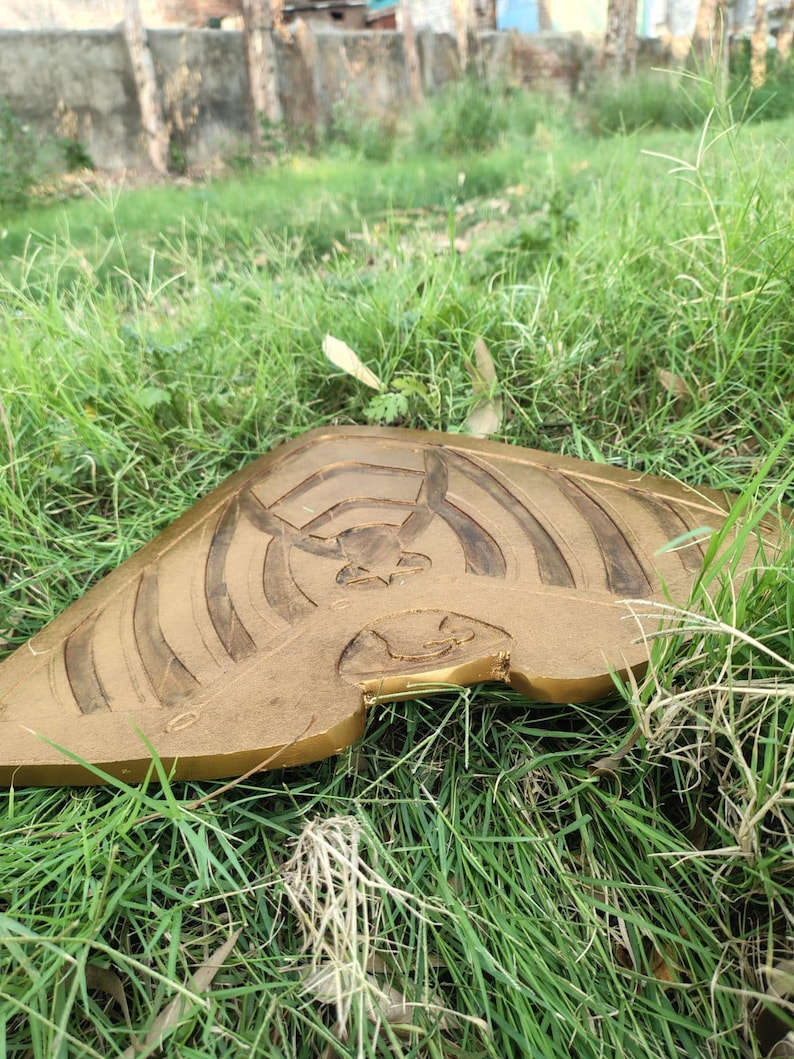 Pode incluir: Um objeto de madeira entalhada de cor dourada com um design estilizado apresentando uma figura central e linhas radiantes. O objeto est&aacute; sobre grama verde, sugerindo um ambiente ao ar livre.