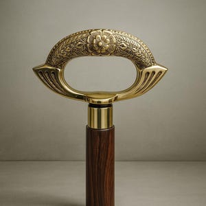 Może przedstawiać: Ozdobna laska do chodzenia z mosiężnym uchwytem i ciemnym drewnianym trzonkiem. Mosiężny uchwyt ma owalny kształt z detalami kwiatowymi i liściastymi. Trzonek wykonany jest z ciemnobrązowego drewna. Klasyczny dodatek.