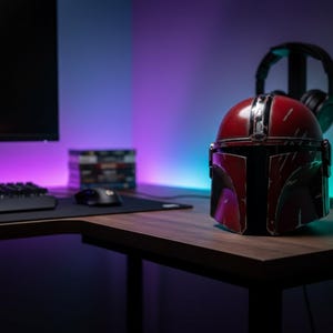 May include: A red and silver Mandalorian helmet sits on a wooden desk. The helmet is the focal point, with a black computer monitor, keyboard, and mouse in the background. A pair of black headphones rests on a stand behind the helmet.