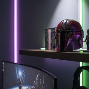 May include: A shelf displays a red and purple Mandalorian helmet, a small figurine, and a stack of DVDs. The helmet is the focal point, with a weathered appearance. The scene is illuminated by purple and green neon lights.