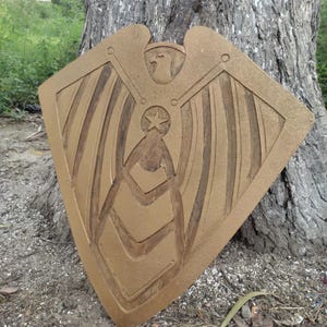 Pode incluir: Um objeto decorativo em forma de escudo, castanho-dourado, com um design de &aacute;guia e estrela. O escudo est&aacute; encostado a um tronco de &aacute;rvore, mostrando detalhes esculpidos de asas e padr&otilde;es geom&eacute;tricos. Parece ser de madeira.