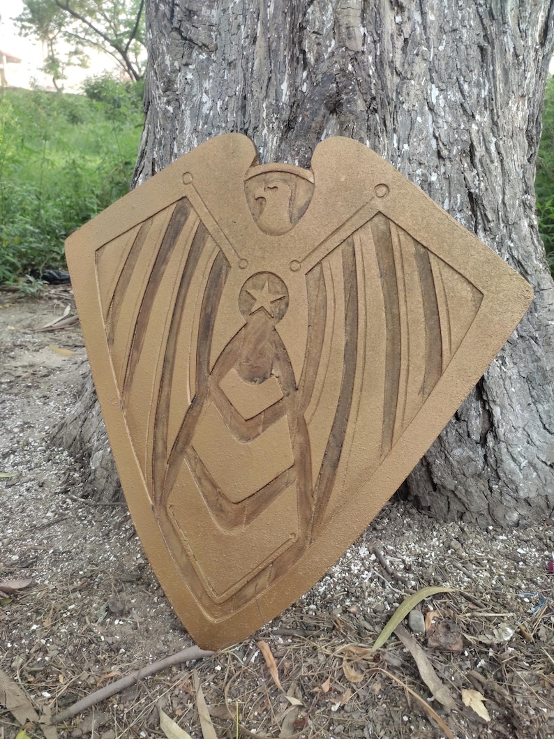 Pode incluir: Um objeto decorativo em forma de escudo, castanho-dourado, com um design de &aacute;guia e estrela. O escudo est&aacute; encostado a um tronco de &aacute;rvore, mostrando detalhes esculpidos de asas e padr&otilde;es geom&eacute;tricos. Parece ser de madeira.