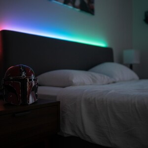 May include: A bedroom scene featuring a bed with white linens and pillows. A Mandalorian helmet sits on a dark nightstand. The headboard has color-changing LED lights. The lights transition from red to blue and green.