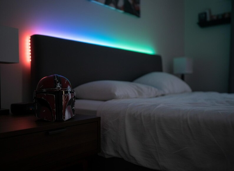 May include: A bedroom scene featuring a bed with white linens and pillows. A Mandalorian helmet sits on a dark nightstand. The headboard has color-changing LED lights. The lights transition from red to blue and green.