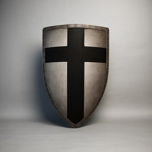 Könnte beinhalten: Ein mittelalterlicher Schild mit silberner Oberfläche und einem markanten schwarzen Kreuz. Der Schild hat eine abgerundete Form mit Metallverkleidung und Nieten an den Kanten. Der Hintergrund ist ein neutrales Grau.