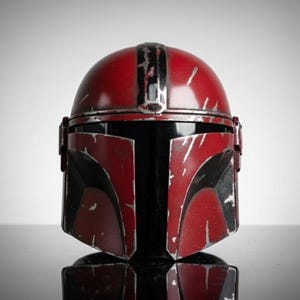 May include: A red and black Mandalorian helmet with a weathered appearance. The helmet features a rounded top with a central stripe and angular cheek plates. The design includes black accents and silver-colored wear marks, giving it a battle-worn look.