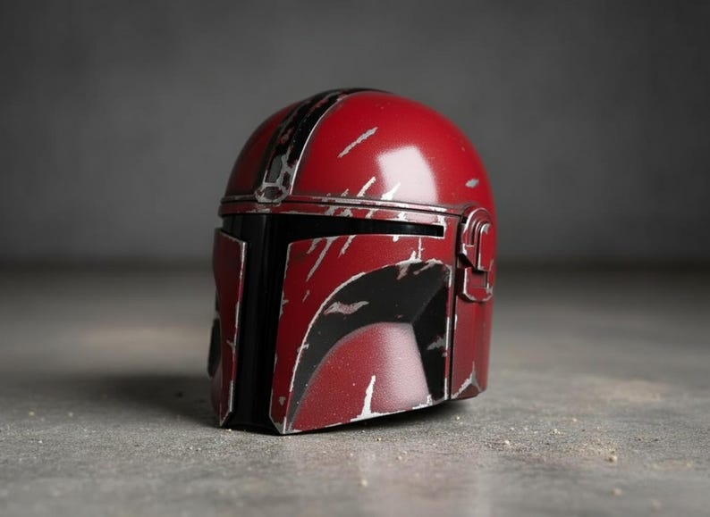 May include: A red and black helmet with silver accents, featuring a weathered, battle-worn appearance. The helmet has a distinctive visor and is likely a prop or collectible item. The design suggests a science fiction or fantasy theme.