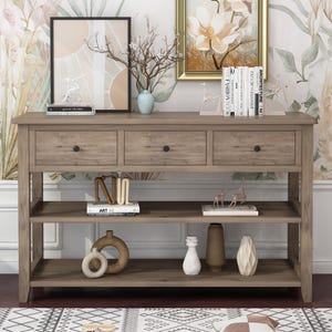 May include: A wooden console table with three drawers and two shelves. The table is a light brown color and has black knobs on the drawers. Decorative items such as vases, books, and artwork are displayed on the shelves and top surface.