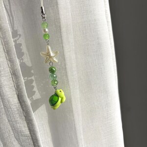 May include: A phone charm featuring a white cord, green and white beads, a white starfish charm, and a small green turtle figurine at the bottom. The accessory is constructed with a series of faceted and smooth beads in various shades of green.