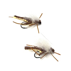 May include: Two artificial fishing flies. Each fly has a brown body with yellow and white accents, a white fluffy wing, and brown legs. A black hook is attached to the bottom of each fly. The flies are set against a white background.