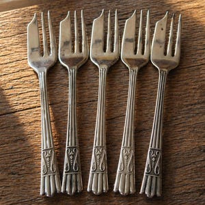 May include: Five vintage silver forks displayed on a wooden surface. The forks feature an ornate design on their handles and are set against a window. The tines of the forks are clearly visible.