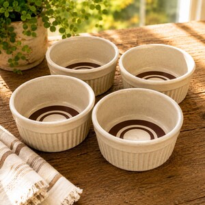 May include: A set of four round, off-white ceramic ramekins with vertical ribbed sides. Each ramekin features a smooth, speckled finish and a dark brown circular ring design centered on the interior base of the dish for a clean, functional look.