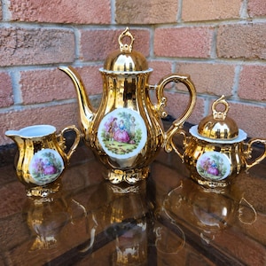 May include: A gold-plated porcelain tea set consisting of a teapot with a lid, a creamer, and a sugar bowl with a lid. Each piece features a central white circular medallion depicting a colourful illustration of a couple in a garden setting.