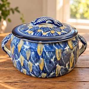 May include: A round ceramic lidded casserole dish featuring a deep blue and white mottled glaze with yellow accents. The lid is topped with a sculpted fish handle, and the sides include two integrated handles shaped like fish heads and tails.