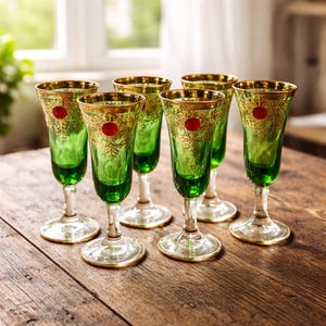 May include: Six green stemmed glasses with gold rims and decorative gold filigree. Each glass has a red dot near the top. The glasses are arranged on a wooden surface, suggesting a vintage or antique aesthetic.