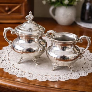 May include: A silver sugar bowl with a lid and a creamer set on a white lace doily. The sugar bowl has a decorative finial, and both pieces have ornate handles and three small feet. The set is reflective and sits on a wooden surface.