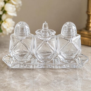 May include: A clear glass salt and pepper set with a matching sugar bowl, presented on a rectangular tray. The set features geometric designs and domed lids. The tray and containers are made of clear glass.