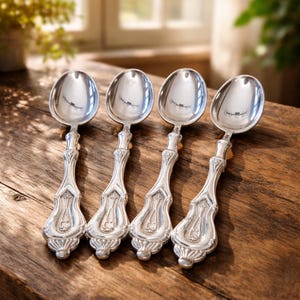 Puede incluir: Cuatro cucharas plateadas con mangos ornamentados se disponen sobre una superficie de madera. Las cucharas tienen un acabado pulido y los mangos presentan un diseño decorativo y vintage. Las cucharas son probablemente para servir o comer.