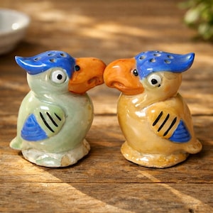 May include: A pair of ceramic parrot-shaped salt and pepper shakers. One is light green with blue accents, the other is yellow with blue accents. Both have orange beaks and blue crests. The shakers are on a wooden surface.