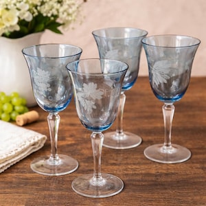Könnte beinhalten: Vier klare Weingläser mit blauen Rändern und Stielen. Jedes Glas hat ein geätztes Blatt- und Traubendesign. Die Gläser stehen auf einer Holzoberfläche, was auf einen Ess- oder Barbereich hindeutet. Im Hintergrund steht eine weiße Vase mit Blumen.