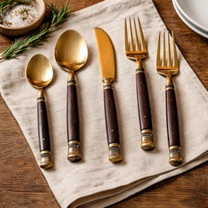 Könnte beinhalten: Ein Set aus fünf goldfarbenen Besteckteilen mit dunkelbraunen Holzgriffen. Das Set umfasst zwei Löffel, ein Messer und zwei Gabeln. Das Besteck ist auf einer beigen Leinen-Serviette angeordnet, mit einer kleinen Schale Salz und Rosmarinzweigen im Hintergrund.