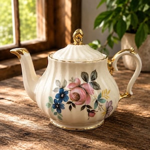 May include: A cream-coloured ceramic teapot with a ribbed texture and gold trim on the handle, spout, and lid finial. The body features a floral pattern with pink roses, blue flowers, and green leaves painted on the front surface of the vessel.